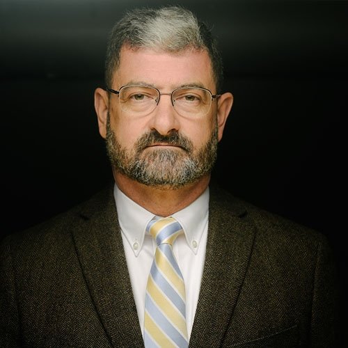 Man with grey hair and beard wearing black suit with gold and silver tie and white dress shirt in front of black background