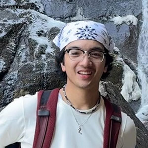Male student wearing bandana, glasses, backpack, white t shirt and necklace in front of outdoor location