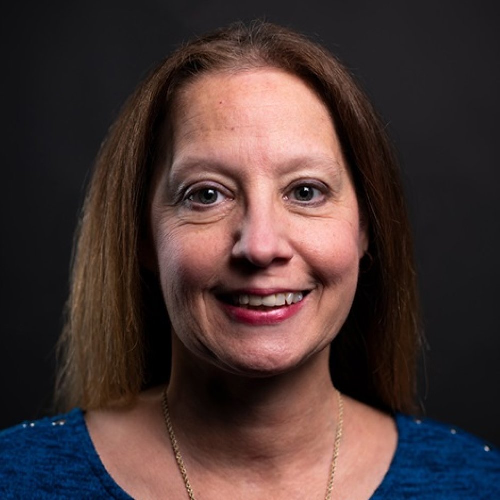 Lady with long dark hair wearing blue top and necklace in front of dark background