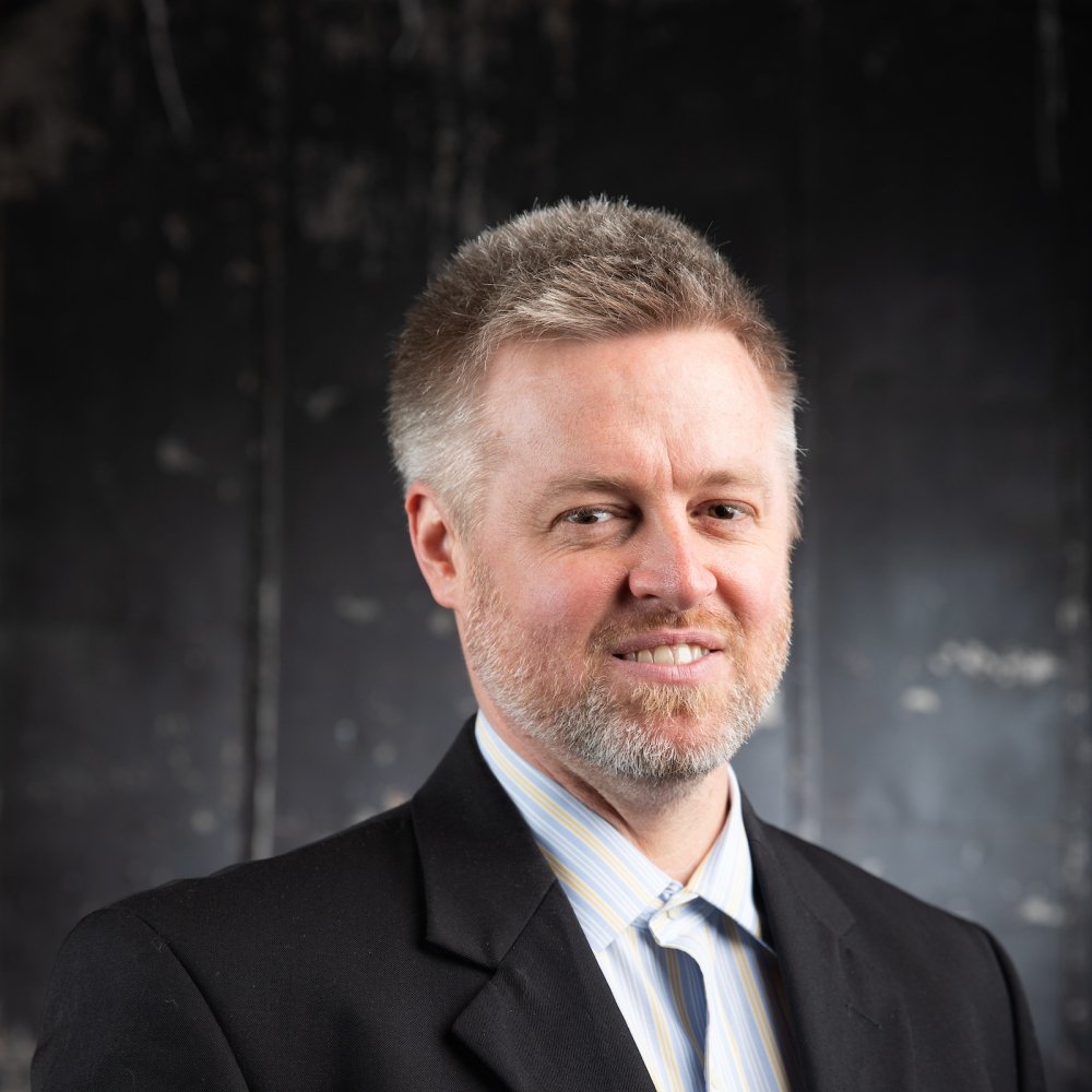 man with dark blonde hair with facial hair wearing dark sports coat, grey button up, and grey tie