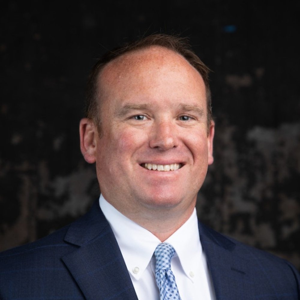 Man with dark suit wearing light blue tie and white button up in front of grey background