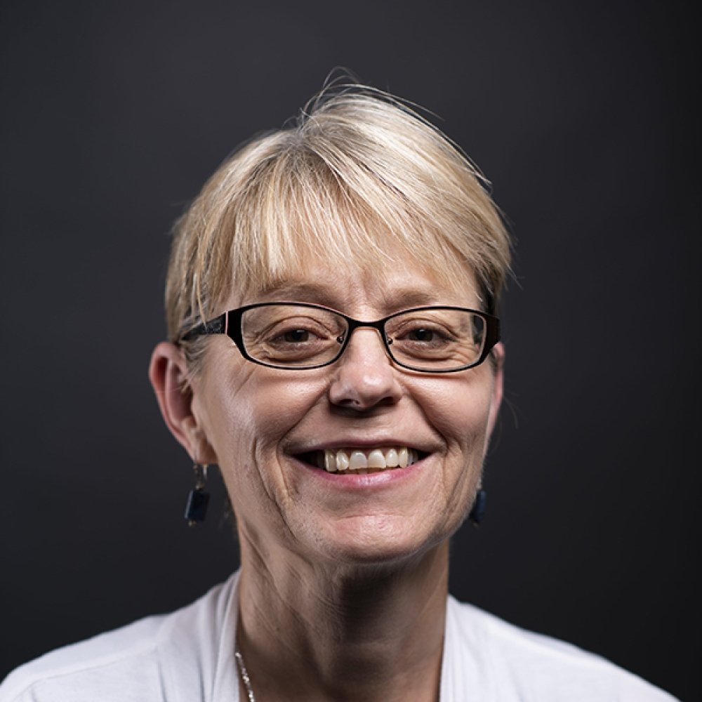 lady with blonde hair pulled back in ponytail wearing glasses with, grey shirt and white cardigan in front of black background