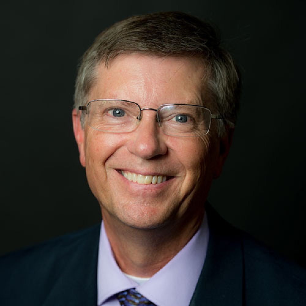 Man with dark hair, glasses, wearing dark suit light purple dress shirt and tie in front of dark background