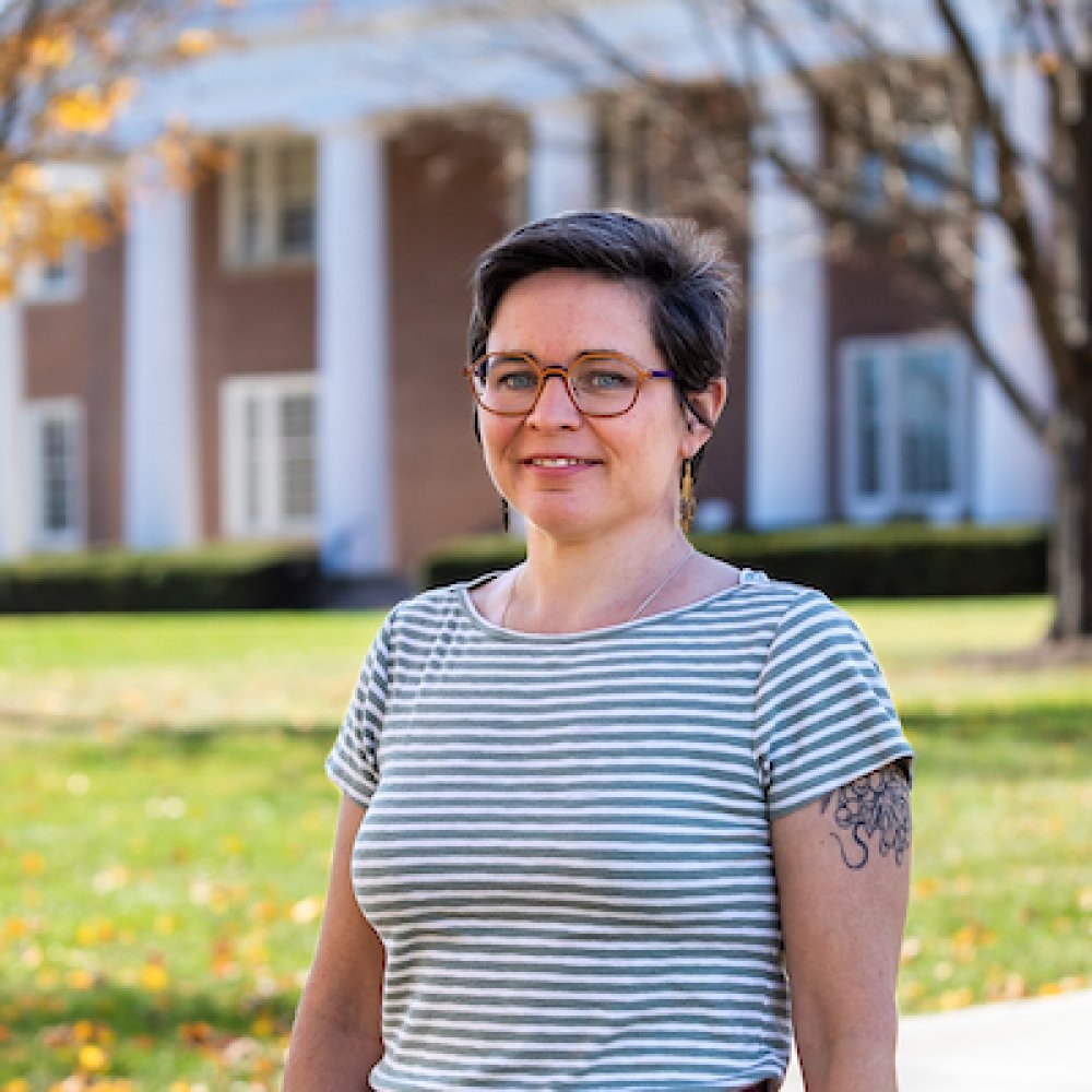 Lady with short dark hair andd glasses, wearing striped short sleeve top standing in front of old centre