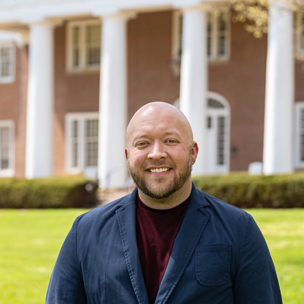 Person with facial hair wearing dark sports coat and black shirt standing in front of Old Centre