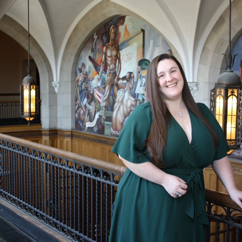 Kasee Arnett  - Lady standing with long brown hair in green dress in historic building next to railing with lights behind her