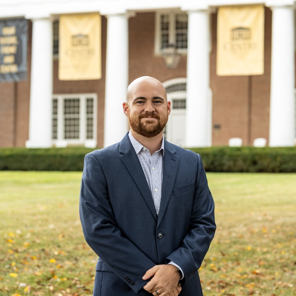 Man with facial hair wearing sports jacket with plaid dress shirt in front of Old Centre