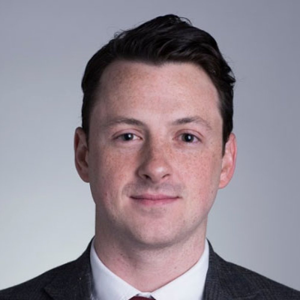 Man with dark hair wearing black suit, white dress shirt, and tie in front of grey background