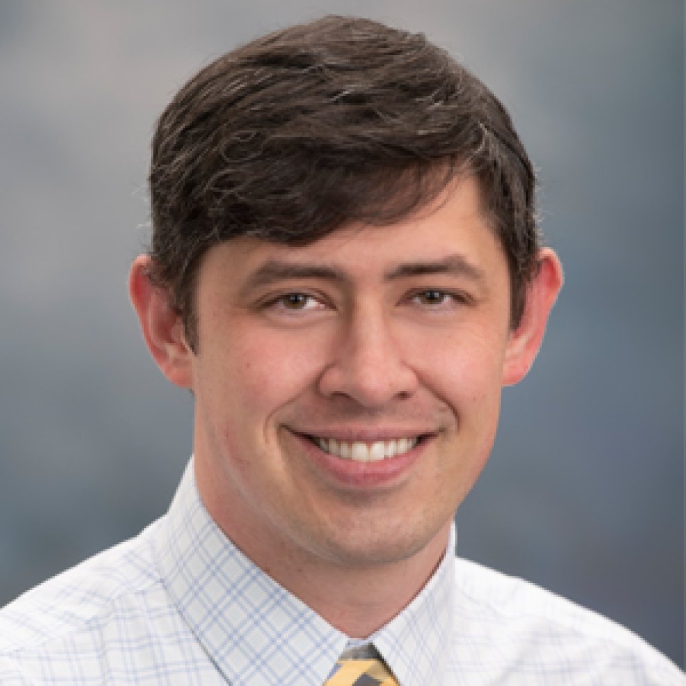 Man with dark hair wearing striped button up dress shirt and yellow tie in front of grey background