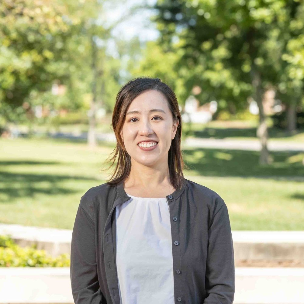 Lady with shoulder length hair wearing black cardigan and white blouse standing outside on campus