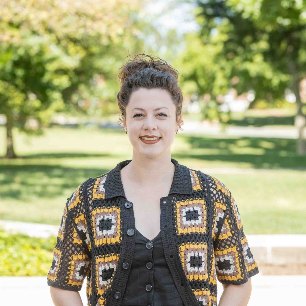 Lady with hair pulled back wearing brown and yellow quilted cardigan and brown top standing outside