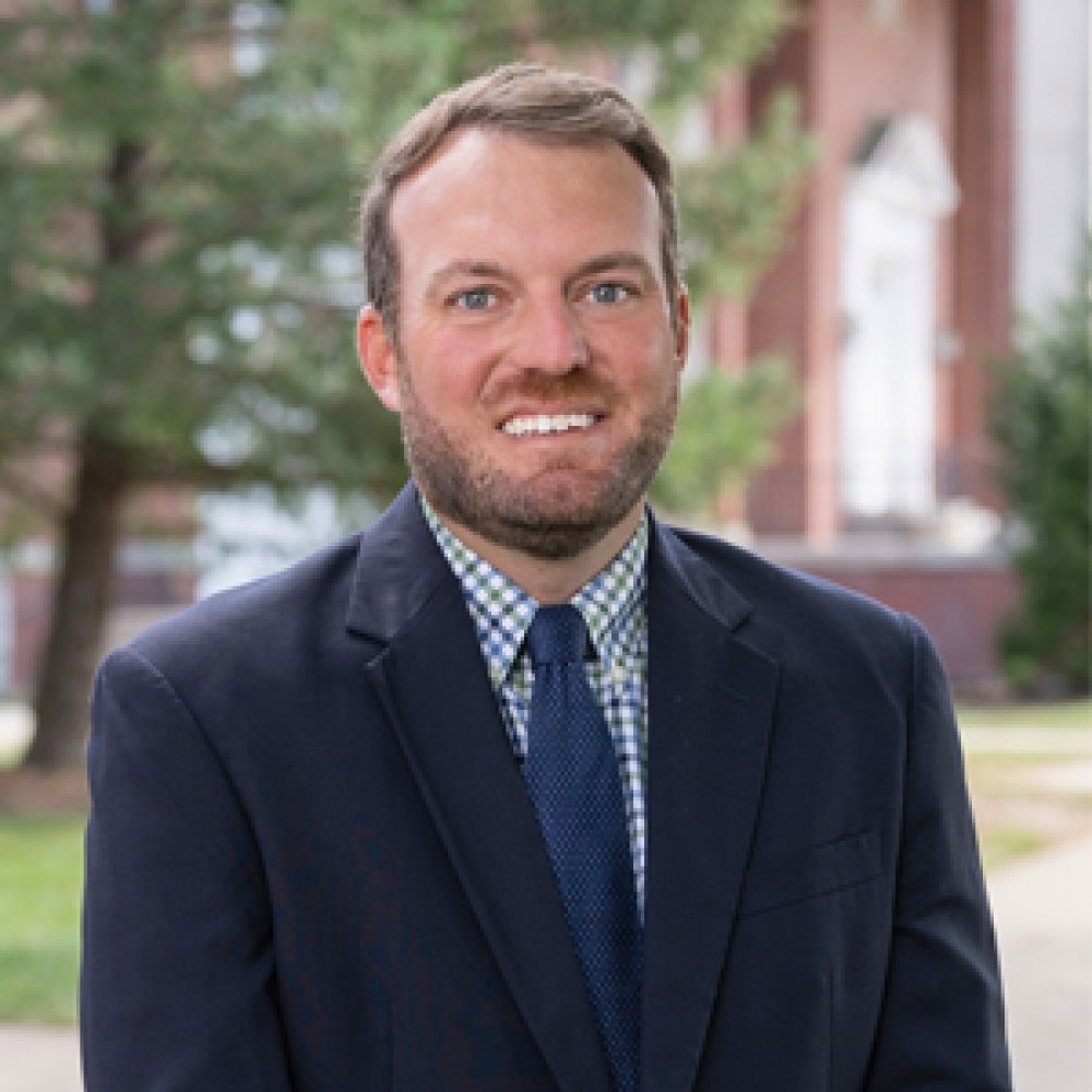 Man with dark hair and beard wearing navy suit with navy tie and gingham dress shirt