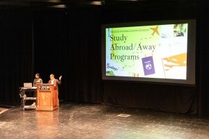 Two people stand behind a podium while a projection screen introduces Centre College Study Abroad &amp; Away programs.
