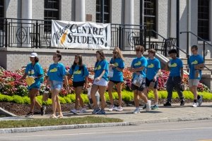 Nine college students wearing matching blue t-shirts pass a banner hanging from a downtown building that states "Welcome Centre Students."