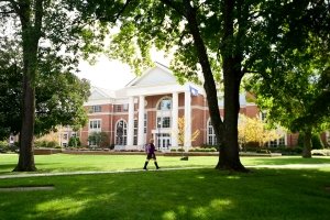 A student walks along a path lined with trees. In the background is Crounse Hall, a large brick building with tall columns.