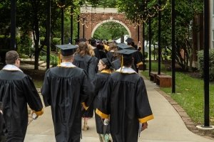 Centre College students wearing commencement robes and caps make their way through a brick archway on campus.