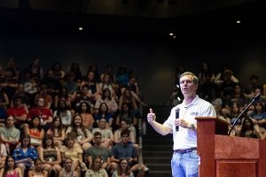 Kentucky Gov. Andy Beshear speaks into a microphone as an auditorium full of high school students look on.