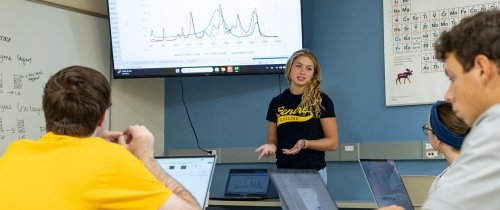 A young woman speaks with follow students seated in a classroom as a line graph is shown on a screen behind her.