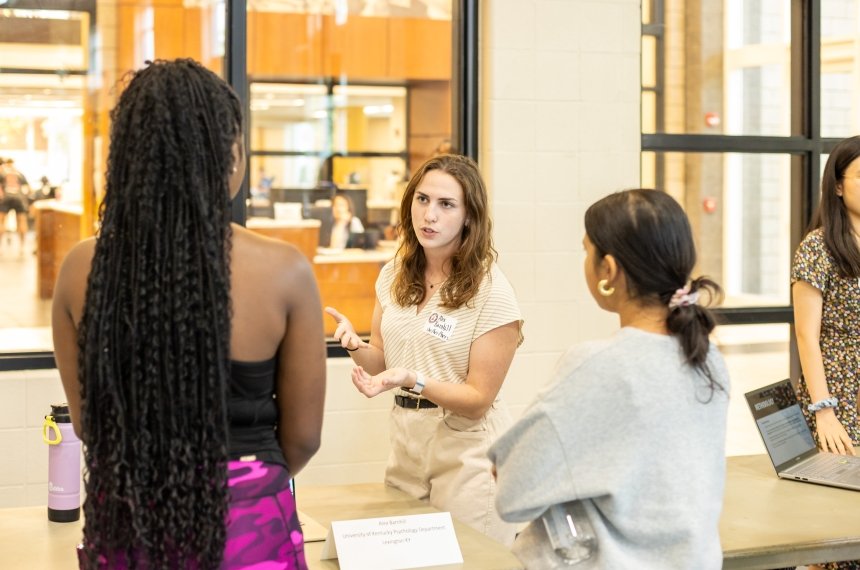 A woman faces the camera as she speaks with two other women standing with their backs to the camera.