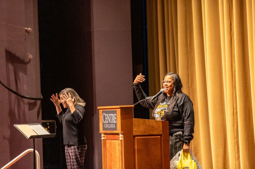 A woman speaks behind a lectern as an interpreter for the deaf and hard of hearing signs for members of the audience.