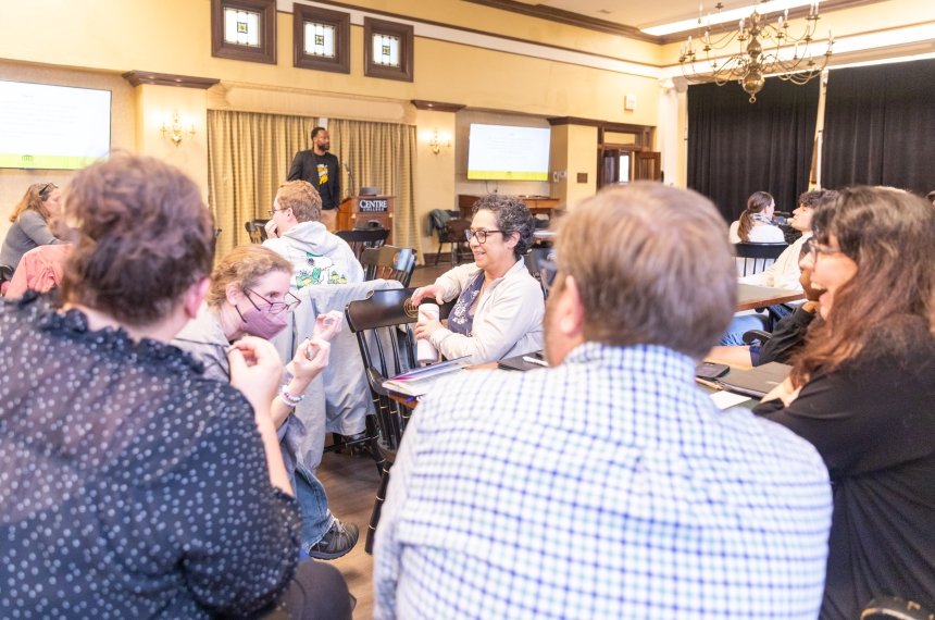 A man stands at the front of a room addresses others seated at tables.