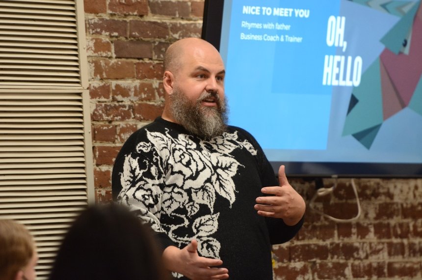 A man stands in front of a screen with the worlds "Oh Hello" on it as she speaks.