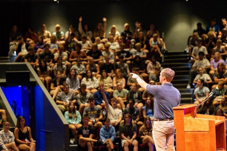 A man points into the audience while speaking from a stage. 