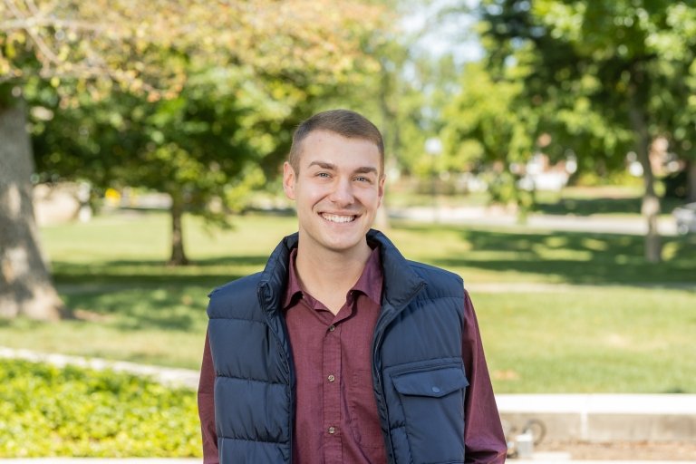 Man with dark hair wearing maroon button up shirt and blue puffy vest