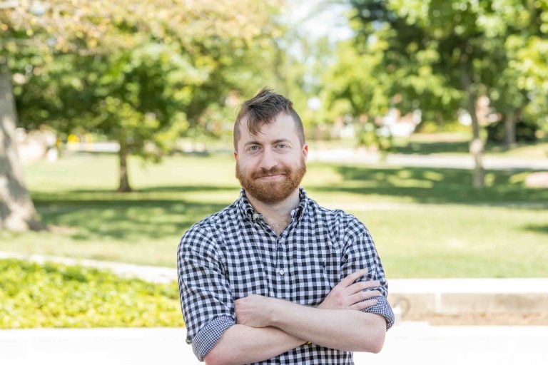 Man with dark hair and beard wearing black and white gingham shirt with arms folded standing outside