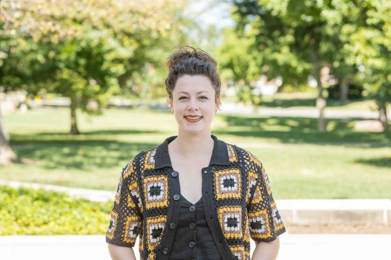 Lady with hair pulled back wearing brown and yellow quilted cardigan and brown top standing outside