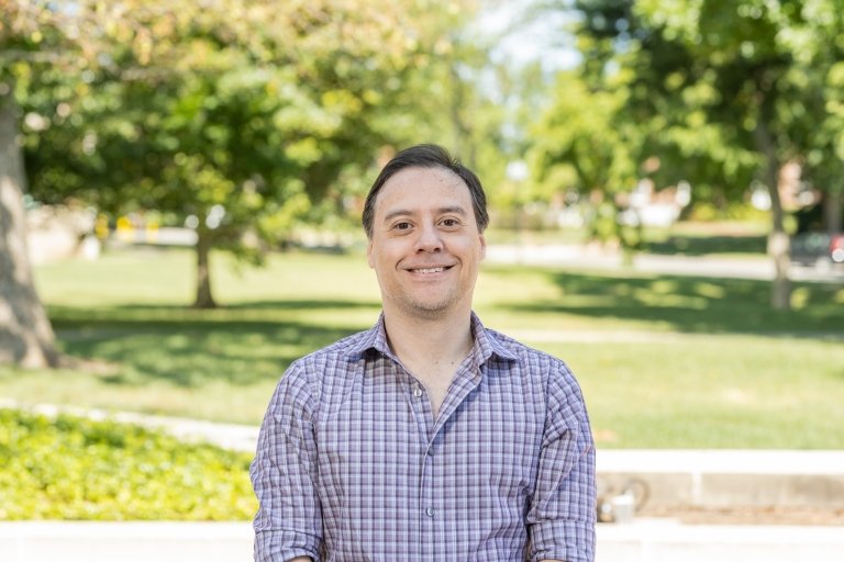 Man with dark hair wearing plaid gingham dress shirt standing outside 