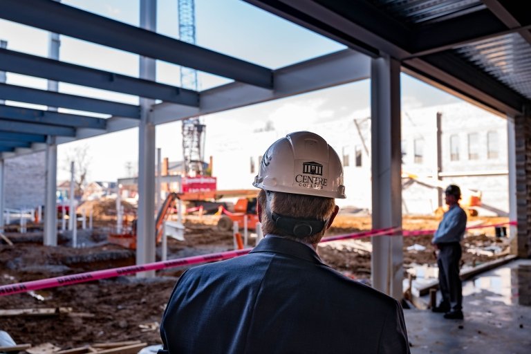 A man in a suit coat and a hardhat looks at the steel framework of an under-construction building. 