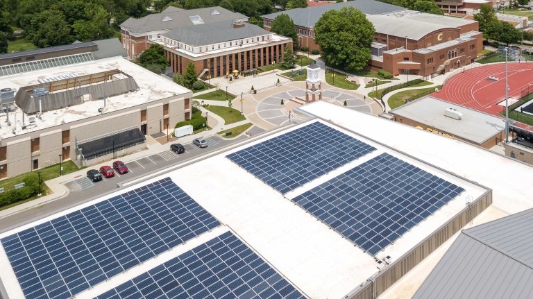 Rows of solar panels are shown on the roof of a building with other buildings visible in the background. 