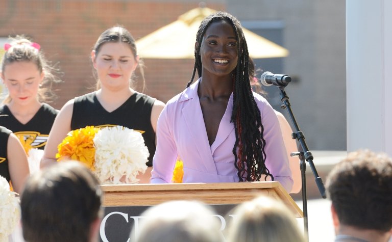 A young woman in a purple jacket speaks into a microphone while standing behind a podium. 