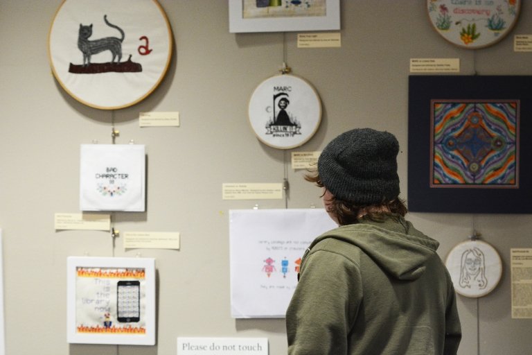 A person is eeen from behind as they look at cross-stitched panels displayed on the wall of Grace Doherty Library. 