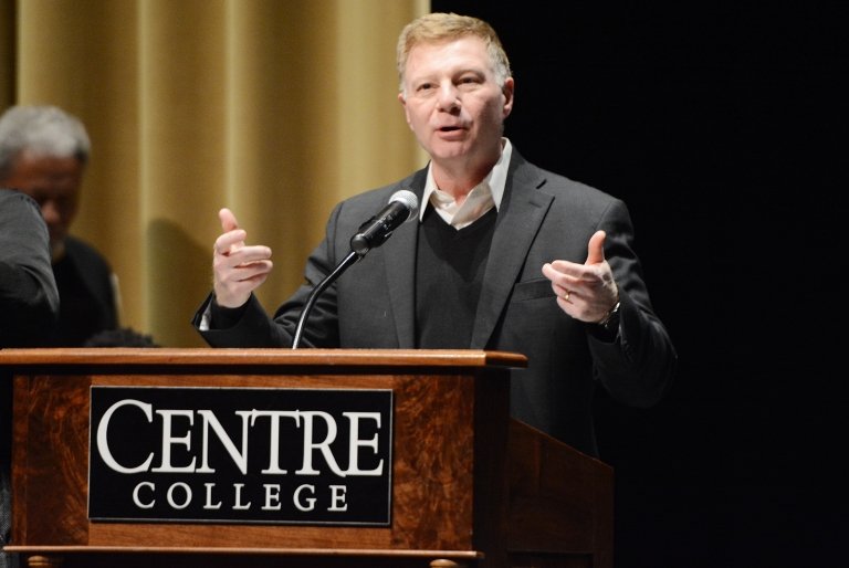 A man speaks into a microphone mounted to a lectern bearing a Centre College sign. 
