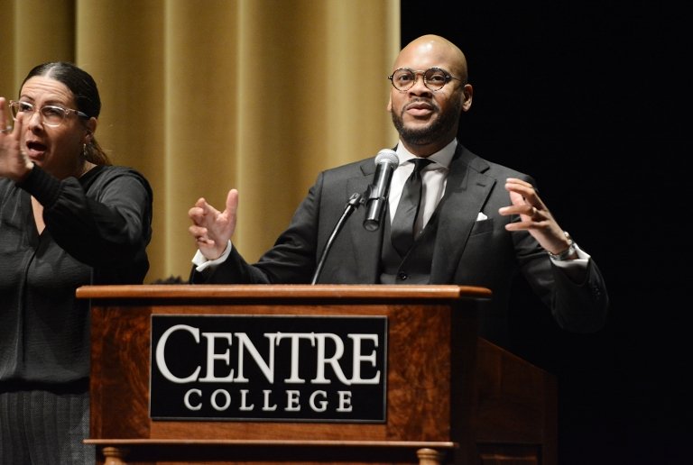 A man speaks into a microphone mounted to a lectern bearing a Centre College sign. 