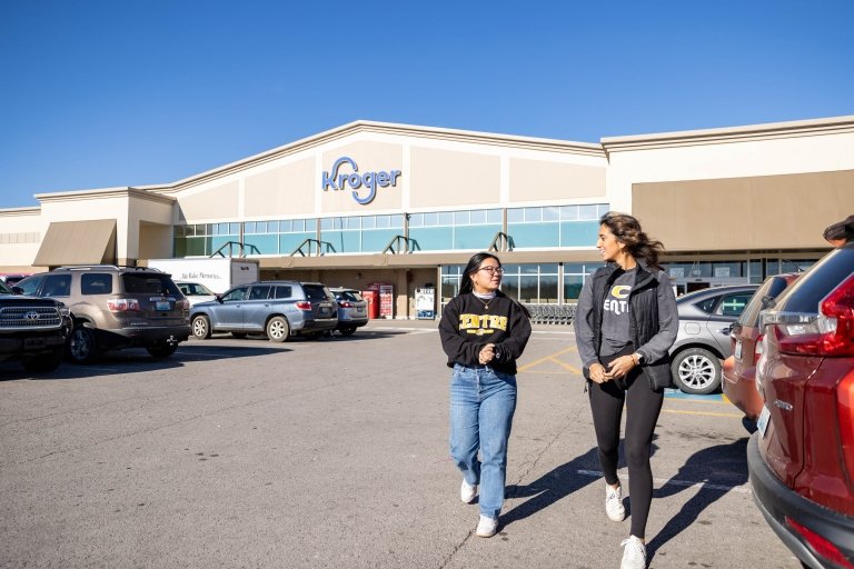 Two women wearing Centre College shirts walk through a parking lot with a Kroger store in the background. 