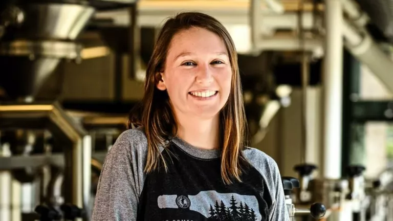 A woman smiles for a photo with large stainless steel tanks and brewing equipment seen in the background. 