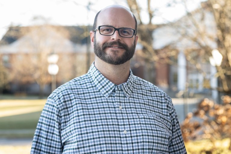 A bearded man wearing black-rimmed glasses and a blue checkered shirt poses for a photo in front of brick buildings. 
