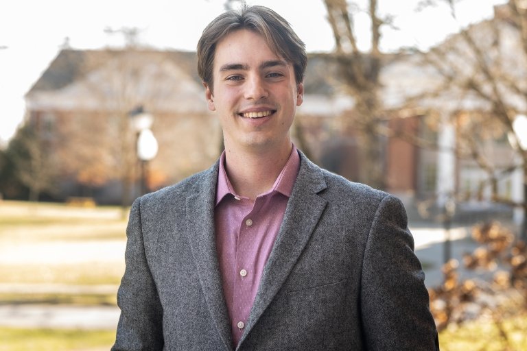 A man wearing a sport coat and purple shirt poses for a photo in front of a brick building with white columns. 