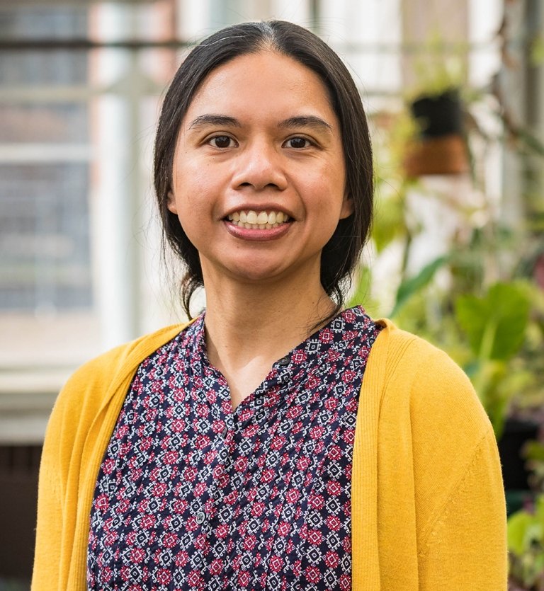 A woman wearing a gold sweat and a colorful shirt poses for a photo in front of a greenhouse full of plants. 