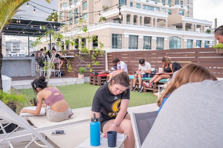 A young woman is among a large group of college students writing on papers outdoors in a city. 