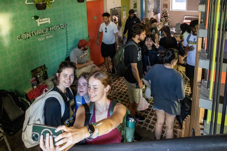 Three women take a selfie in front of a group of other students. 