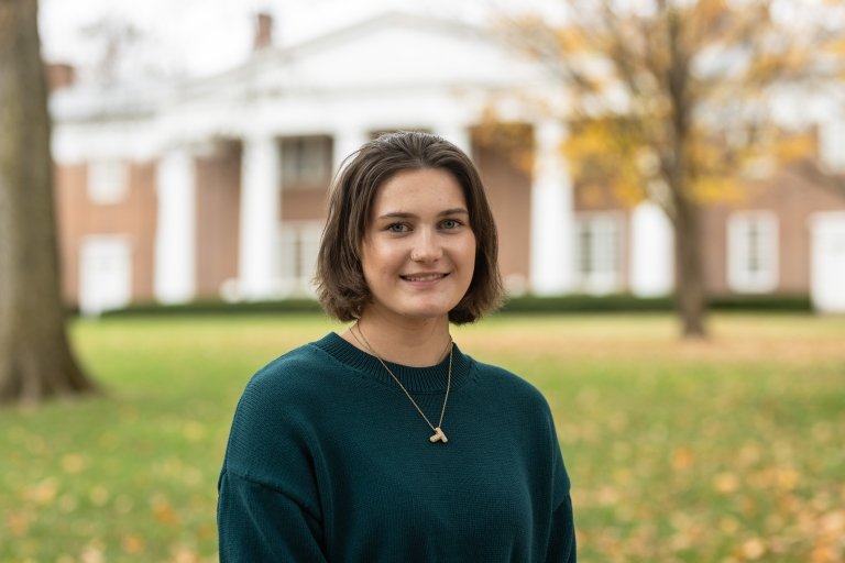 A college-aged woman poses for a photo with a brick building in the background. 