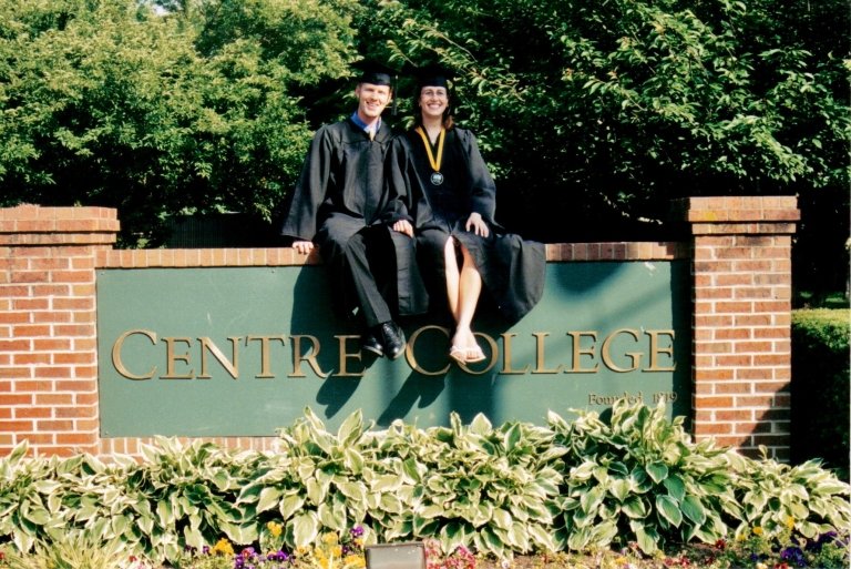 A man and woman dressed in black cap and gown sit atop of brick sign that reads "Centre College" as they celebrate their college graduation. 