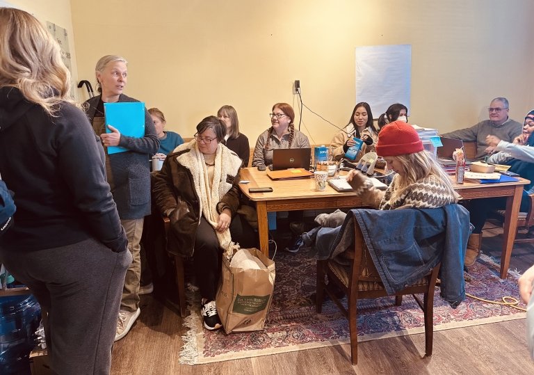Marsha Mount Shoop, holding a bright blue file folder, addresses a group of volunteers, most of them seated around wooden tables filled with computers and papers. 