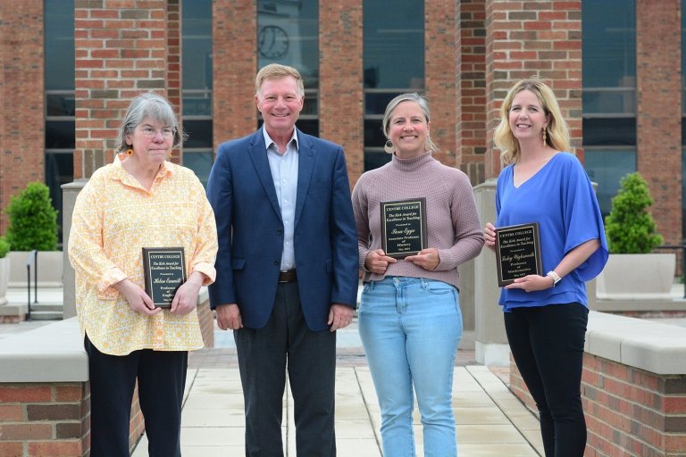 Three women  pose with Centre President Milton C. Moreland for a group photo in front of a brick building while holding certificates of recognition. 