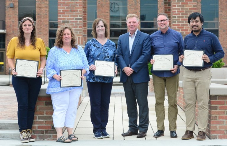 Three women and two men pose with Centre President Milton C. Moreland for a group photo in front of a brick building while holding certificates of recognition. 
