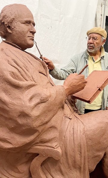A man wearing a yellow beret uses a clay sculpting tool to shape the face of a seated figure he is creating in a studio space. 